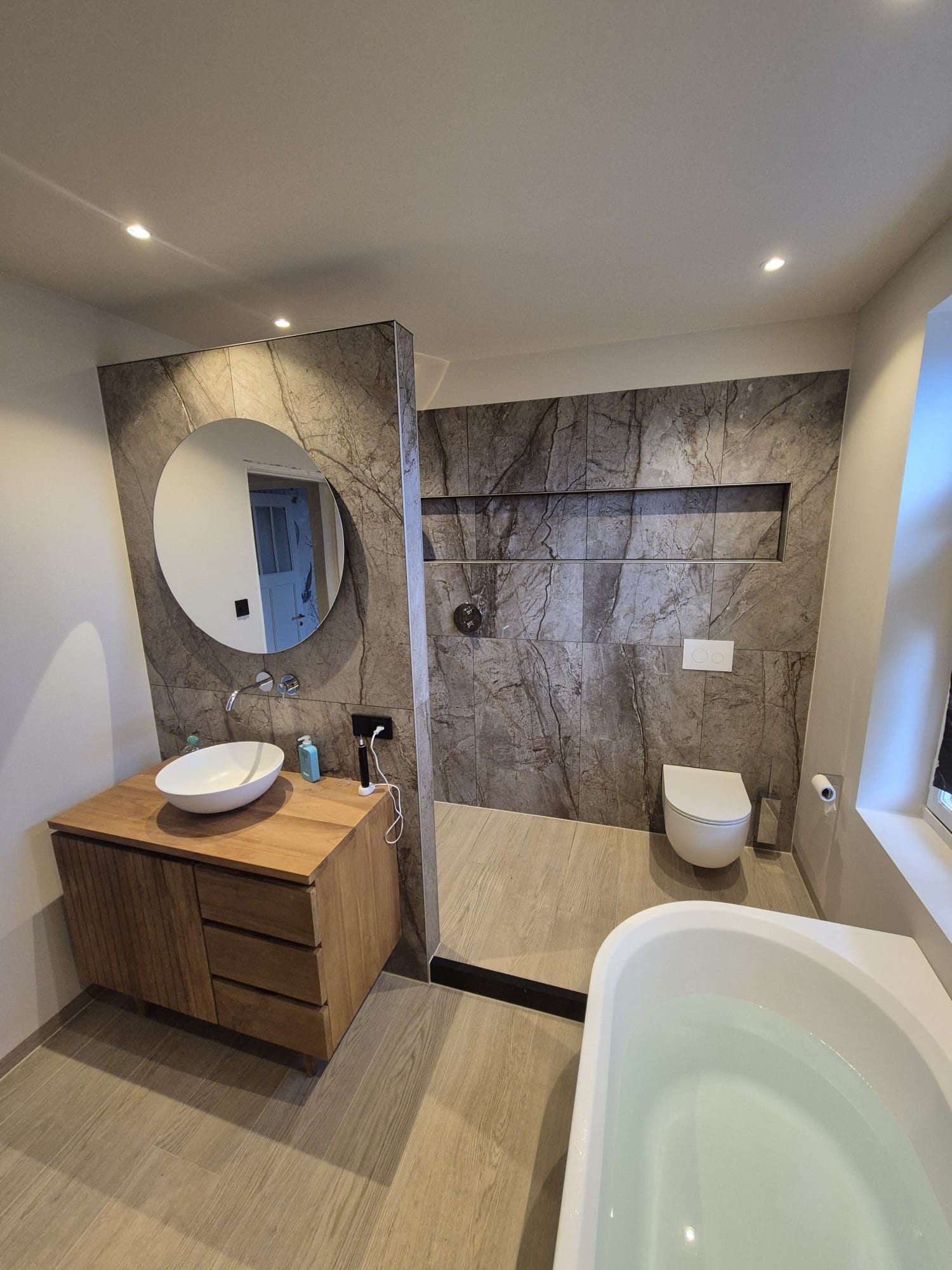 Badkamer met natuursteenlook wand, ronde spiegel, houten wastafelmeubel en vrijstaand bad